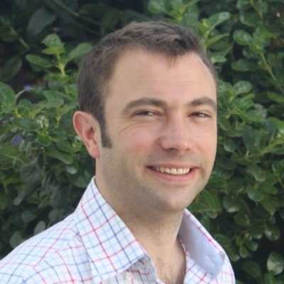 Chris Kelly - A person in a checkered shirt smiles in front of a leafy green background.