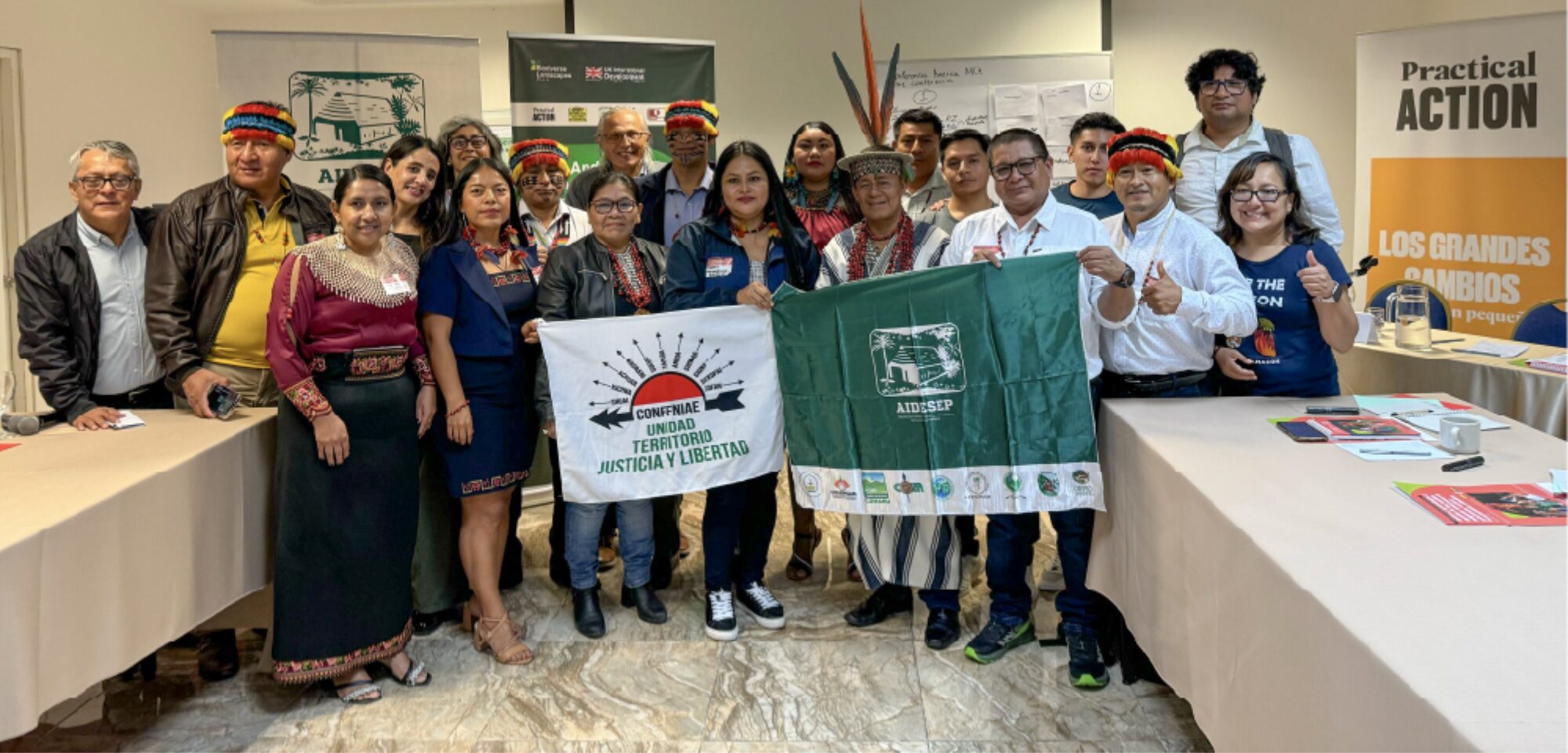A group of people pose indoors, some wearing traditional clothing and headdresses, holding banners with organizational logos and slogans. Tables with pamphlets and banners are in the background.