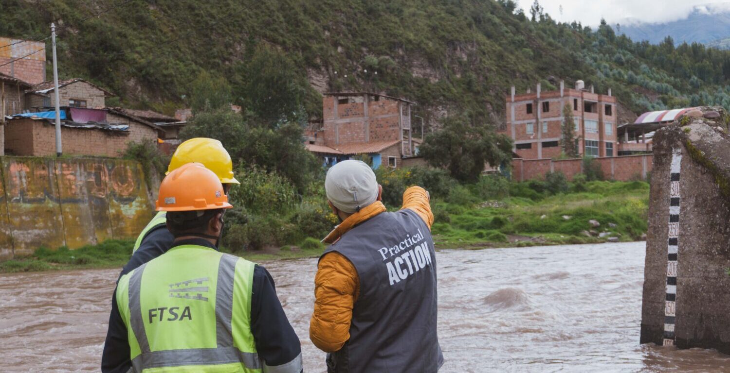Three people wearing safety gear stand by a river, with one person pointing towards buildings across the water in a rural area.
