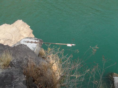 A long metal pipe extends horizontally from a rocky cliff over turquoise water, secured by concrete at the edge.