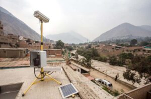 Air quality monitoring device with a solar panel installed on a rooftop, overlooking a residential area with mountains in the background under hazy skies.
