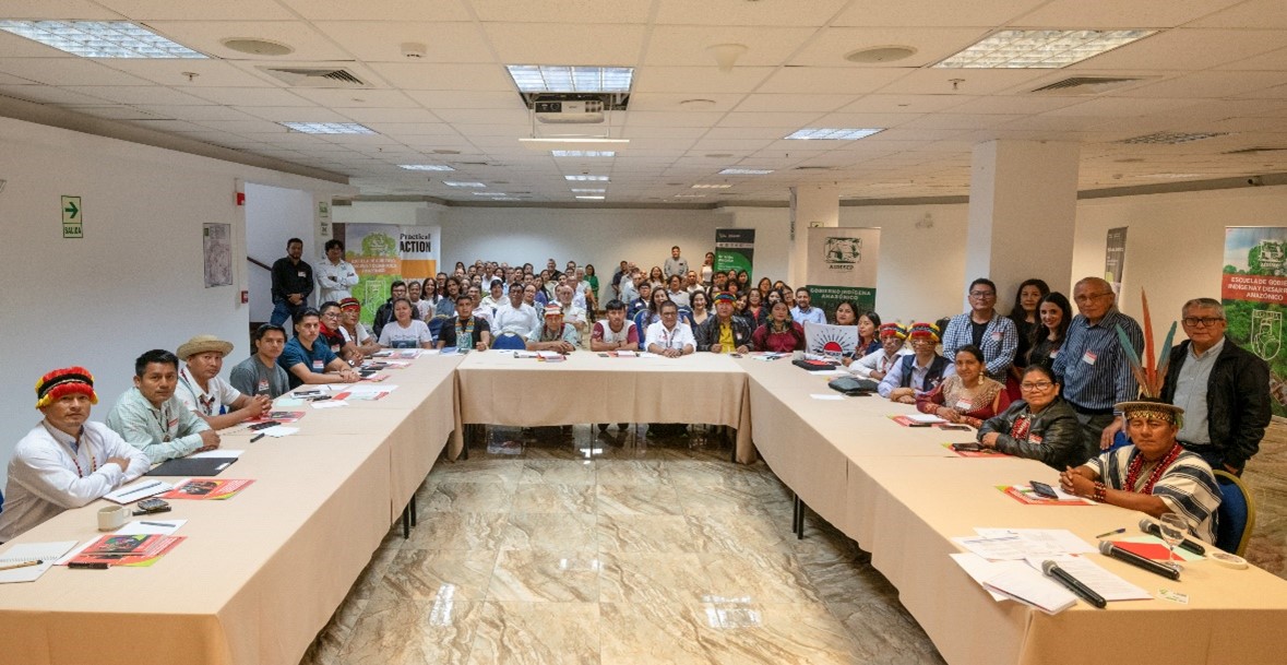 A large group of people sits and stands around a U-shaped conference table in a meeting room, facing the camera.