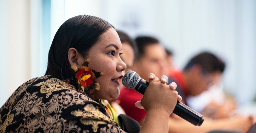 A woman with floral earrings speaks into a microphone during a meeting, with other people seated in the blurred background.