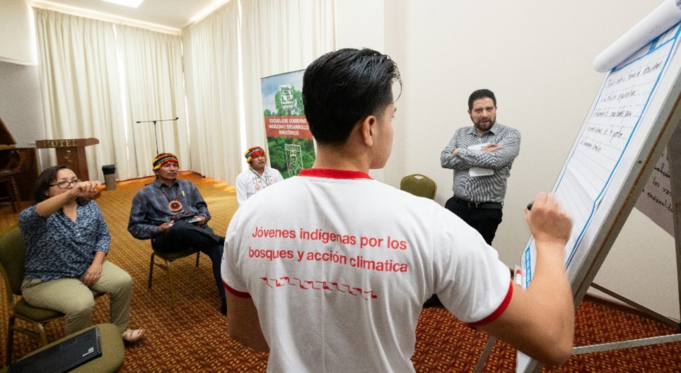 A man writes on a flip chart in a small meeting room as several people, including Indigenous youth, sit and listen. His shirt reads,