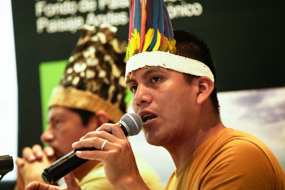Two men wearing feathered headdresses speak into microphones at an event, with a dark background displaying partially visible text.