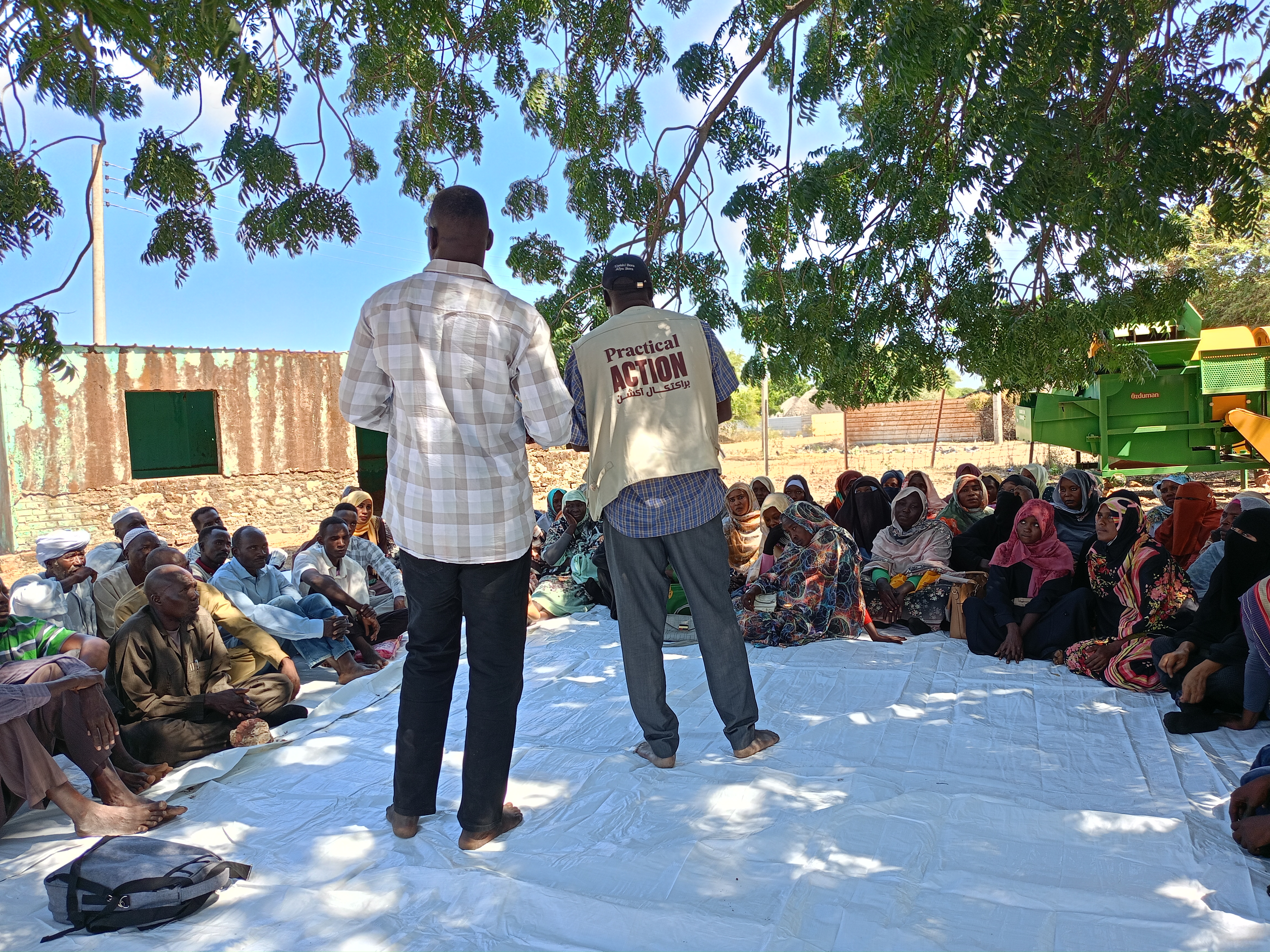 Two men stand and address a seated group of people outdoors under a tree, with one man wearing a vest labeled 