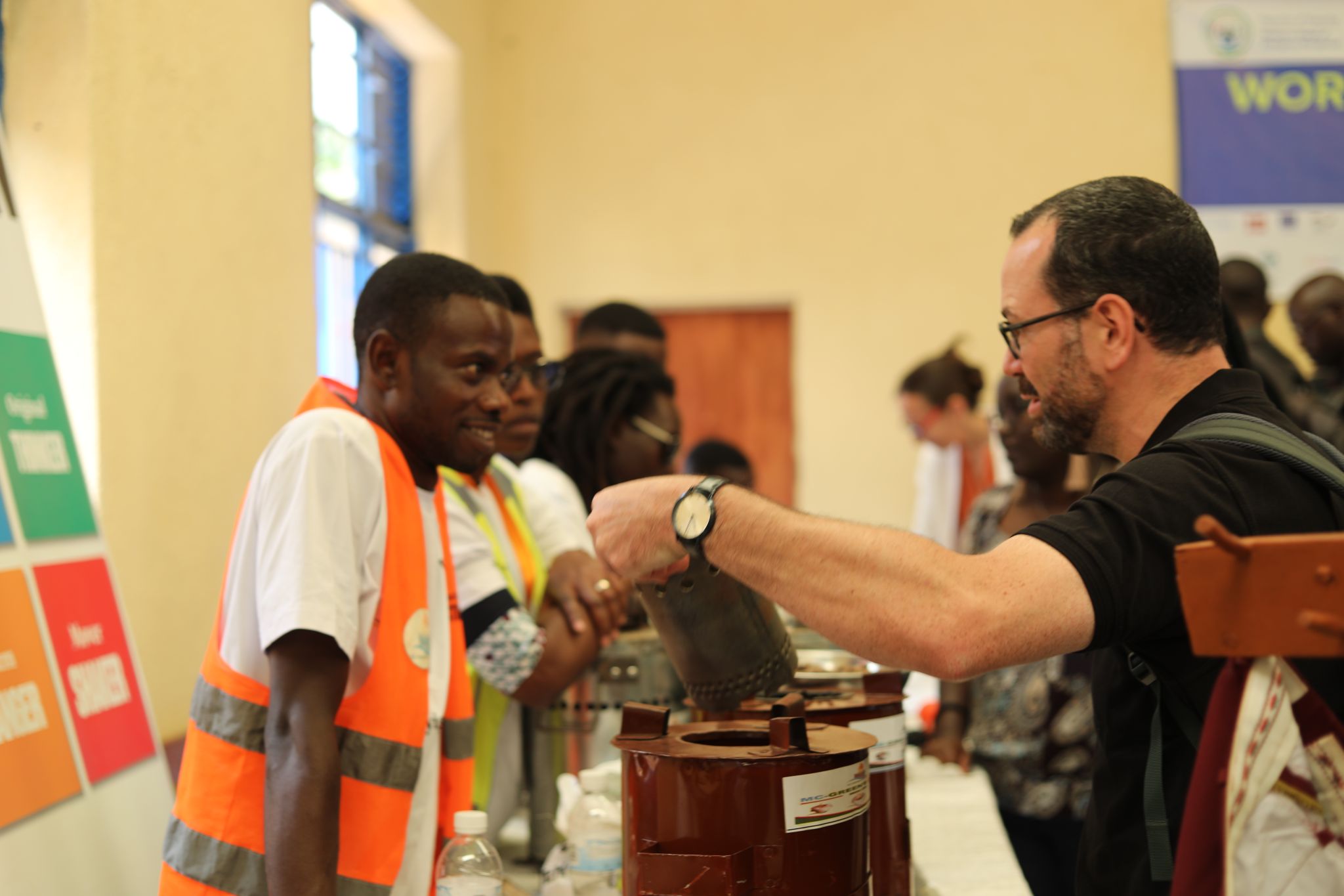A man in a safety vest speaks with another man demonstrating a device at an indoor event with several people and informational signs in the background.