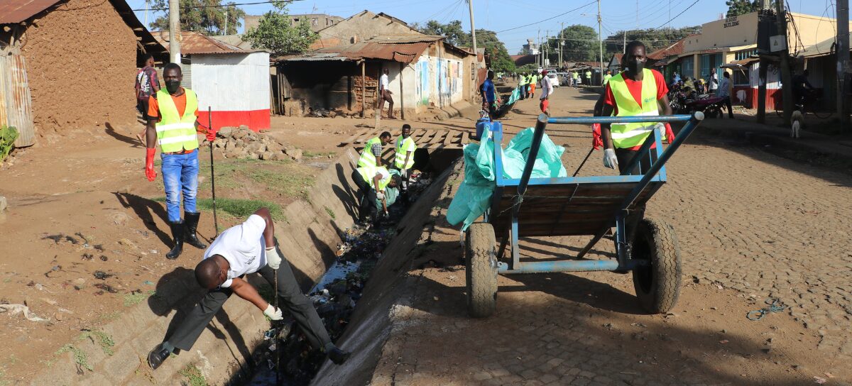 Several people wearing reflective vests clean a roadside drainage ditch and collect debris in bags and a handcart on a cobblestone street in an urban area.