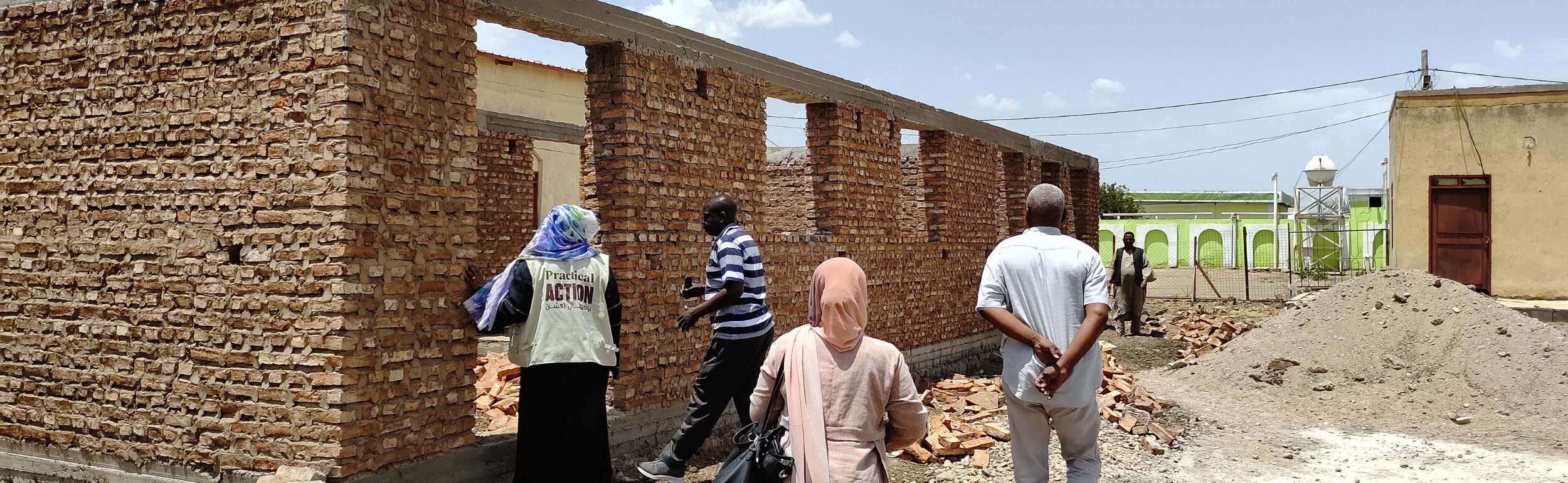 Several people stand beside a partially constructed brick building under a clear sky, with construction materials and debris on the ground.