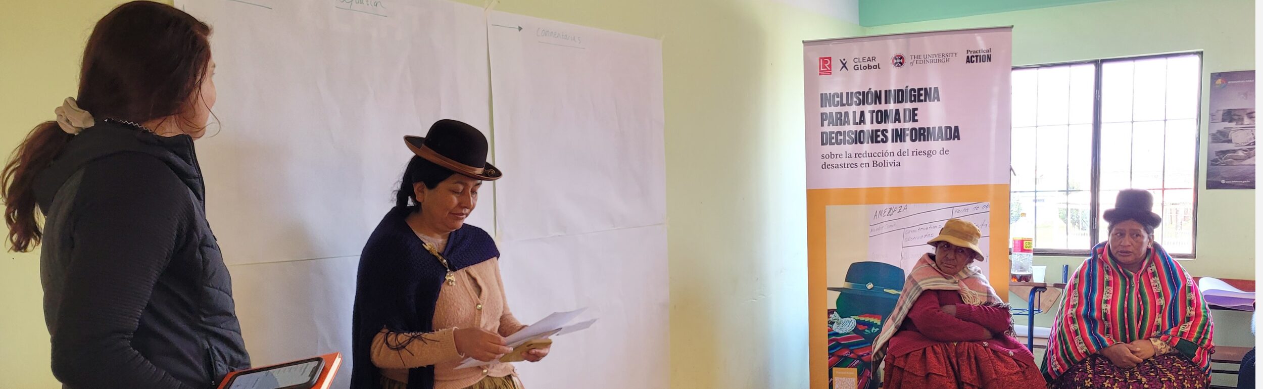A woman stands reading from a paper beside another woman, while two women sit nearby in traditional clothing; a banner about adolescent girls’ education is displayed behind them.