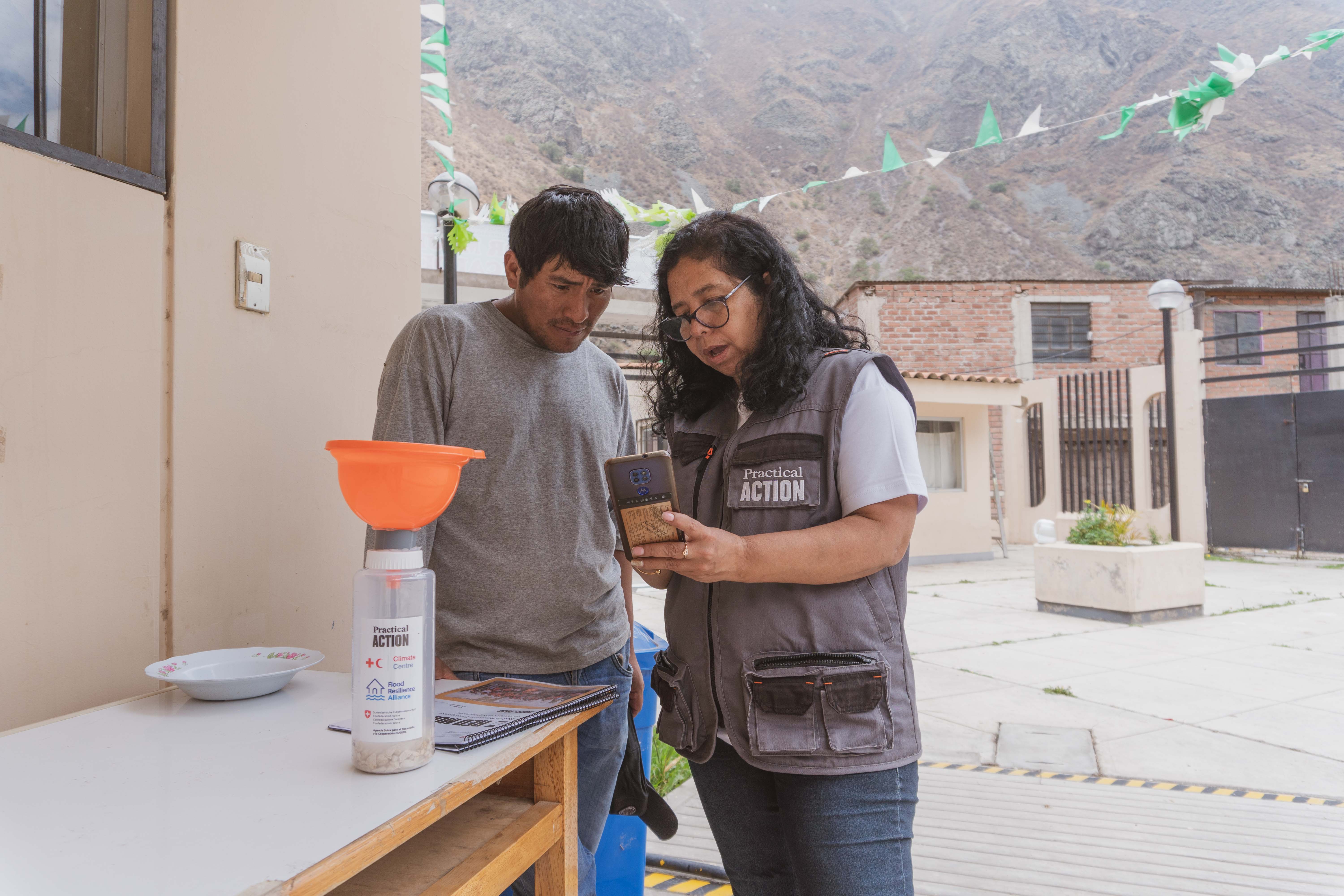 A woman in a Practical Action vest shows a phone to a man outdoors, with a table, funnel, and buildings in the background.