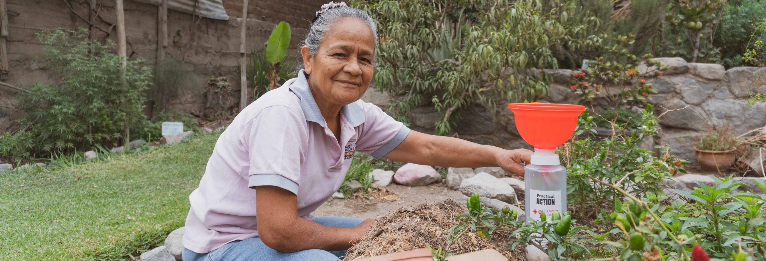 An older woman kneels in a garden, holding a plastic bottle with a red funnel, surrounded by plants and greenery.