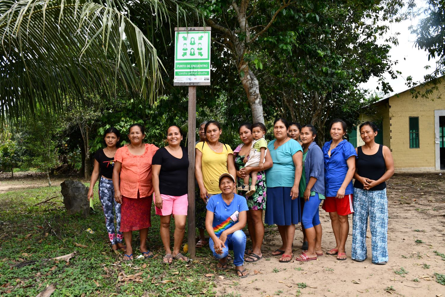 A group of women stands together outdoors in front of trees and a building, beneath a sign on a post.