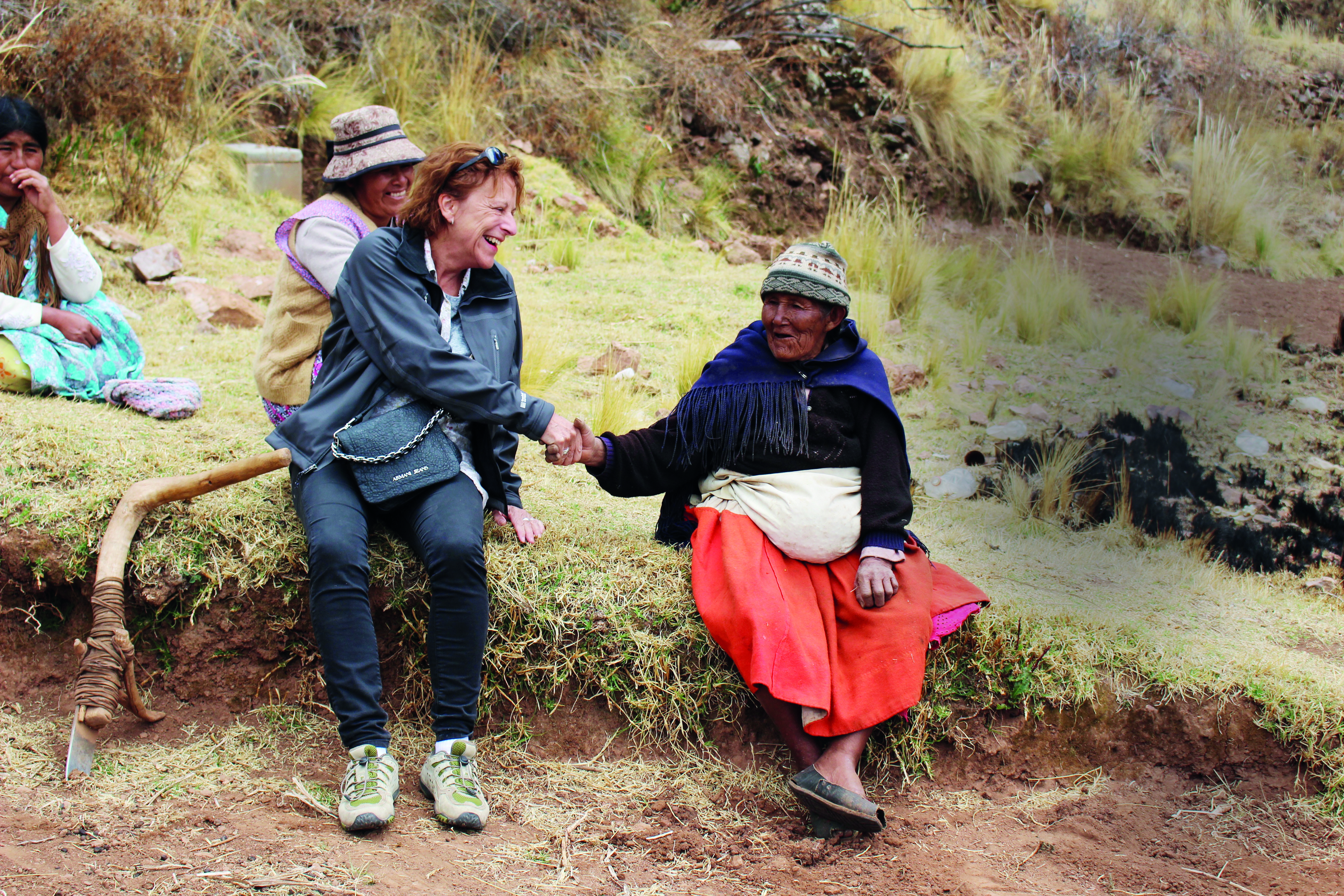 A woman in outdoor clothing shakes hands and smiles with an elderly woman in traditional attire, sitting together on a grassy area with others in the background.