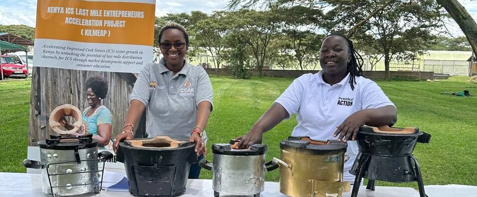 Two women stand behind a table displaying various cookstoves outdoors, with an informational poster and trees in the background.