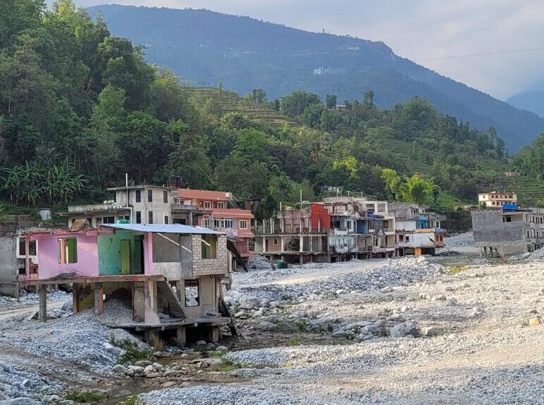 A row of colorful houses stands on rocky terrain beside a dry riverbed, with green hills and cloudy sky in the background.