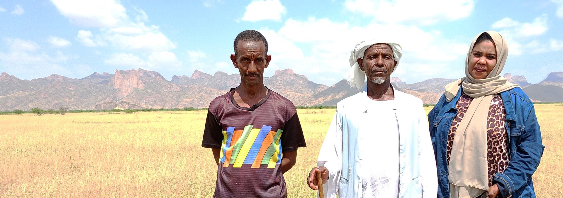 Three people stand in a grassy field with mountains in the background under a partly cloudy sky.