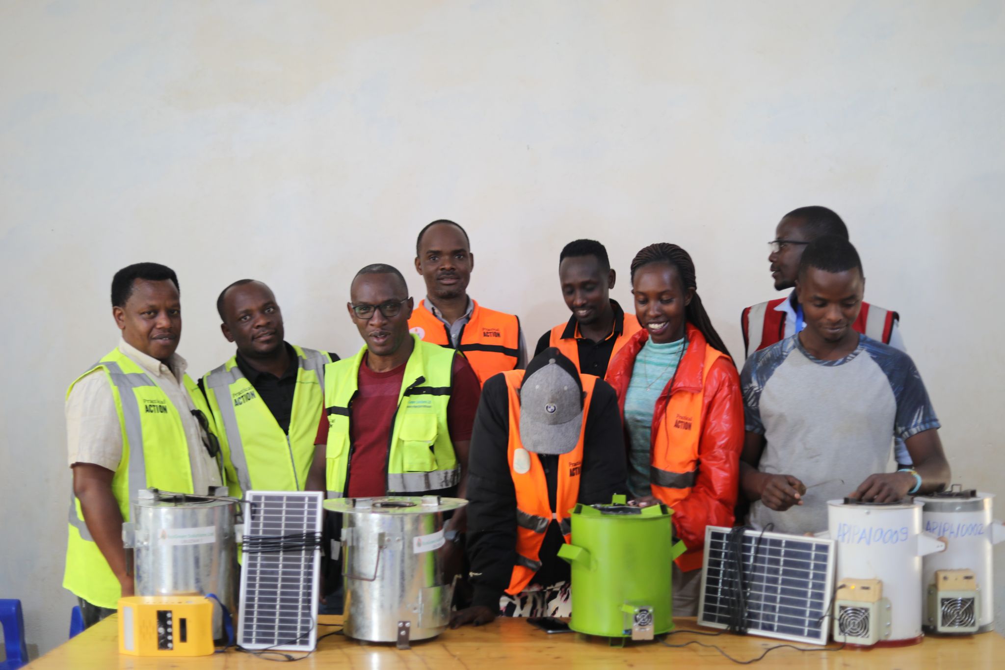 A group of people in safety vests stand behind a table displaying portable stoves and solar panels against a plain background.