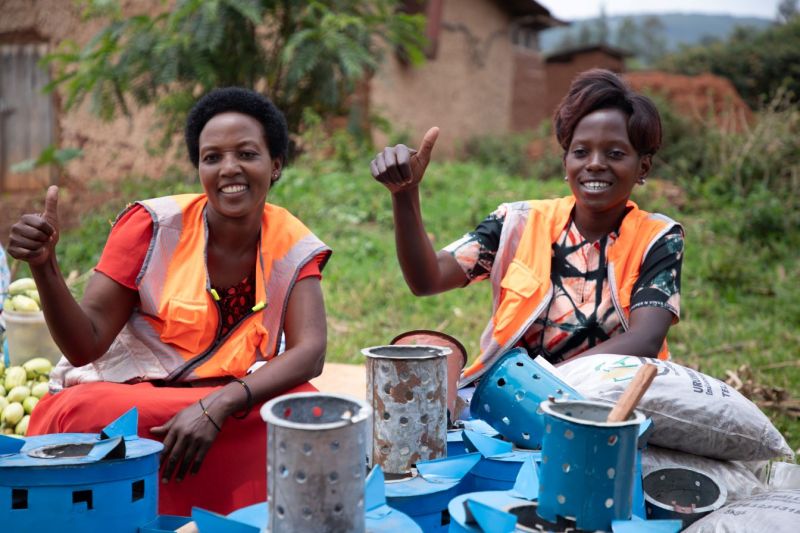 Two women wearing orange safety vests sit outdoors, smiling and giving thumbs up, with metal stoves and bags around them.