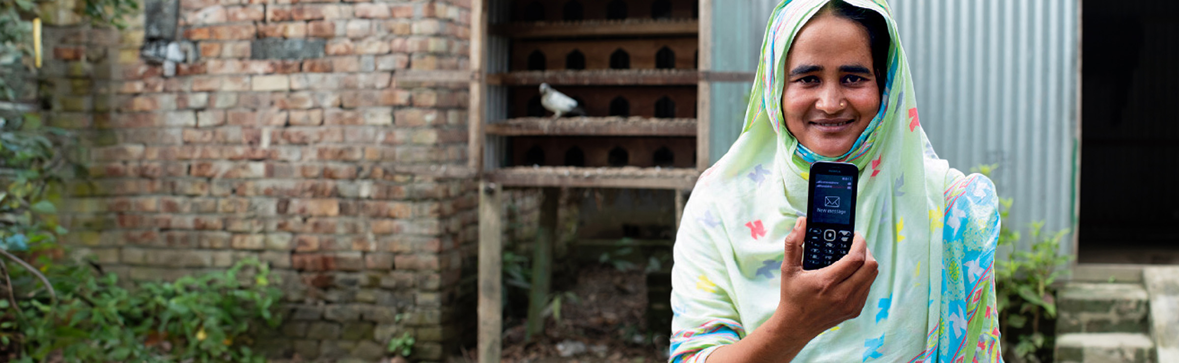 A woman in a patterned headscarf stands outside a brick building, smiling and holding up a mobile phone displaying a text message.