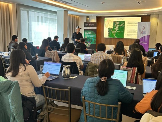 People attend a workshop in a conference room, facing a presenter and a large screen displaying slides and maps; most attendees have laptops open.
