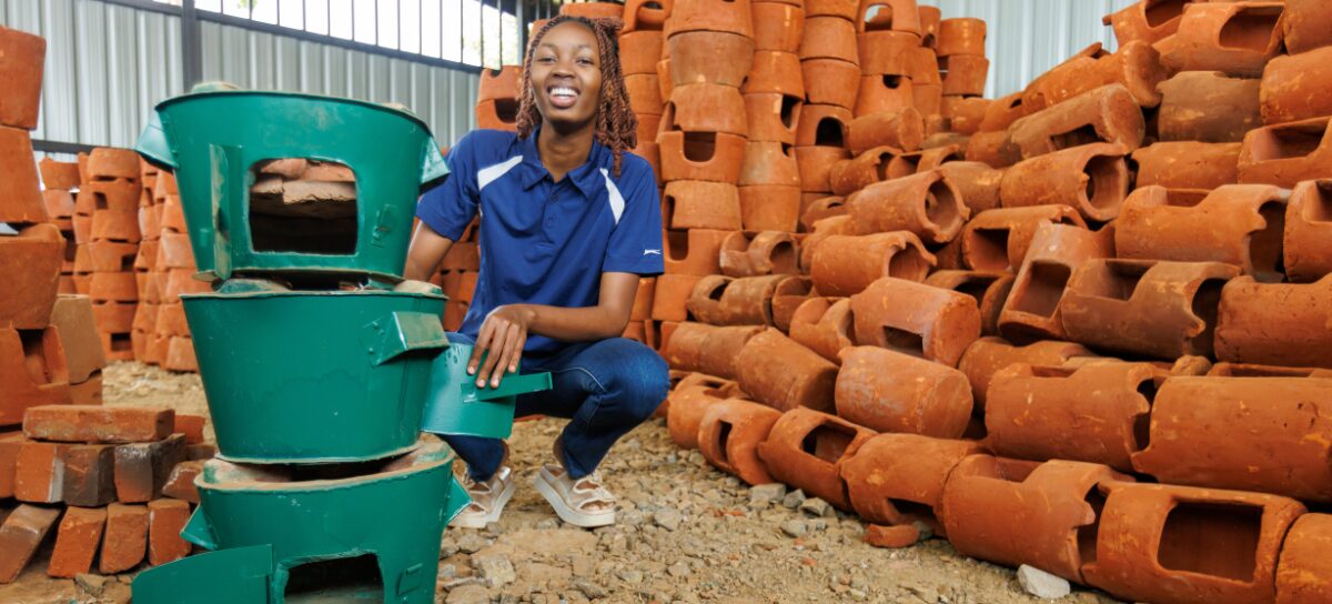 A person squats next to a stack of green stoves, surrounded by piles of reddish-brown clay stove housings in a warehouse setting.