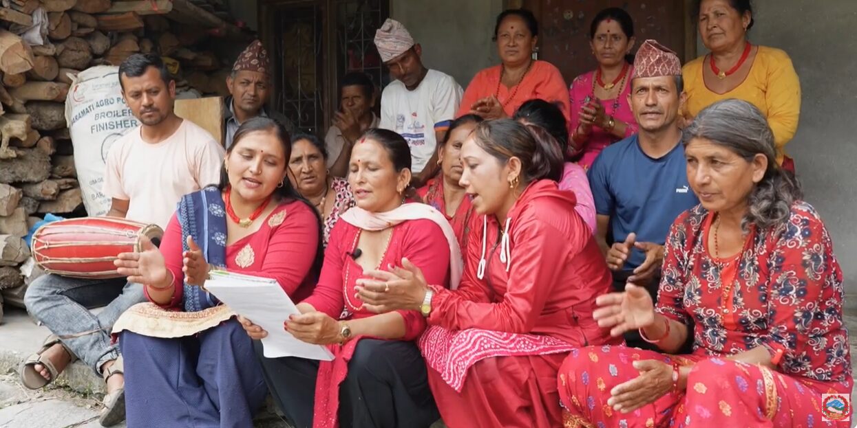 A group of people, mostly women in traditional Nepali attire, sit together singing and clapping, with one person playing a drum and others holding a sheet of paper.