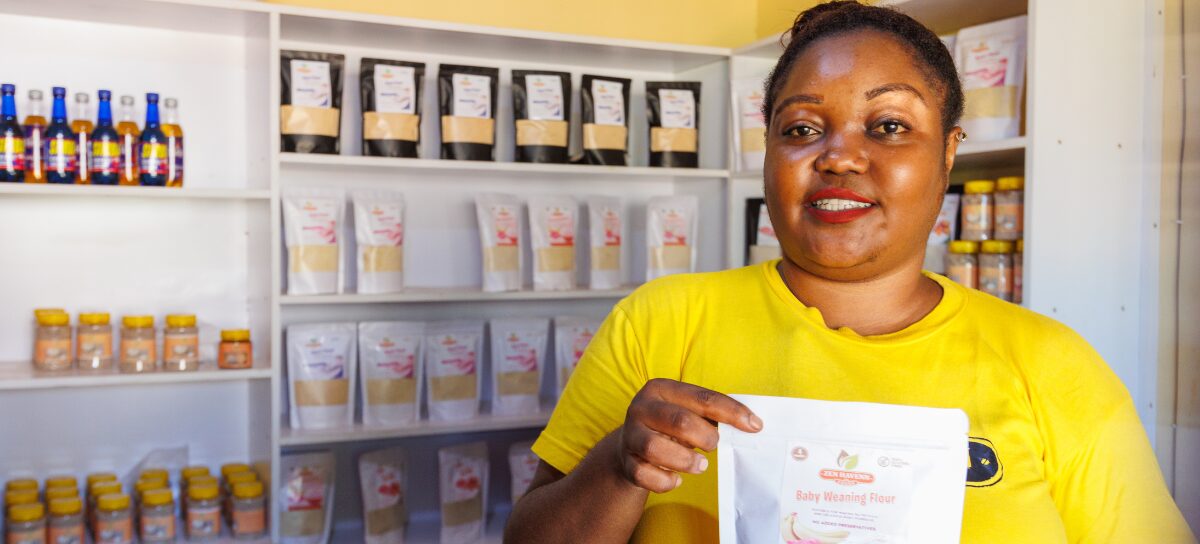 A woman in a yellow shirt holds a package of baby weaning flour in a shop with shelves of food products in the background.