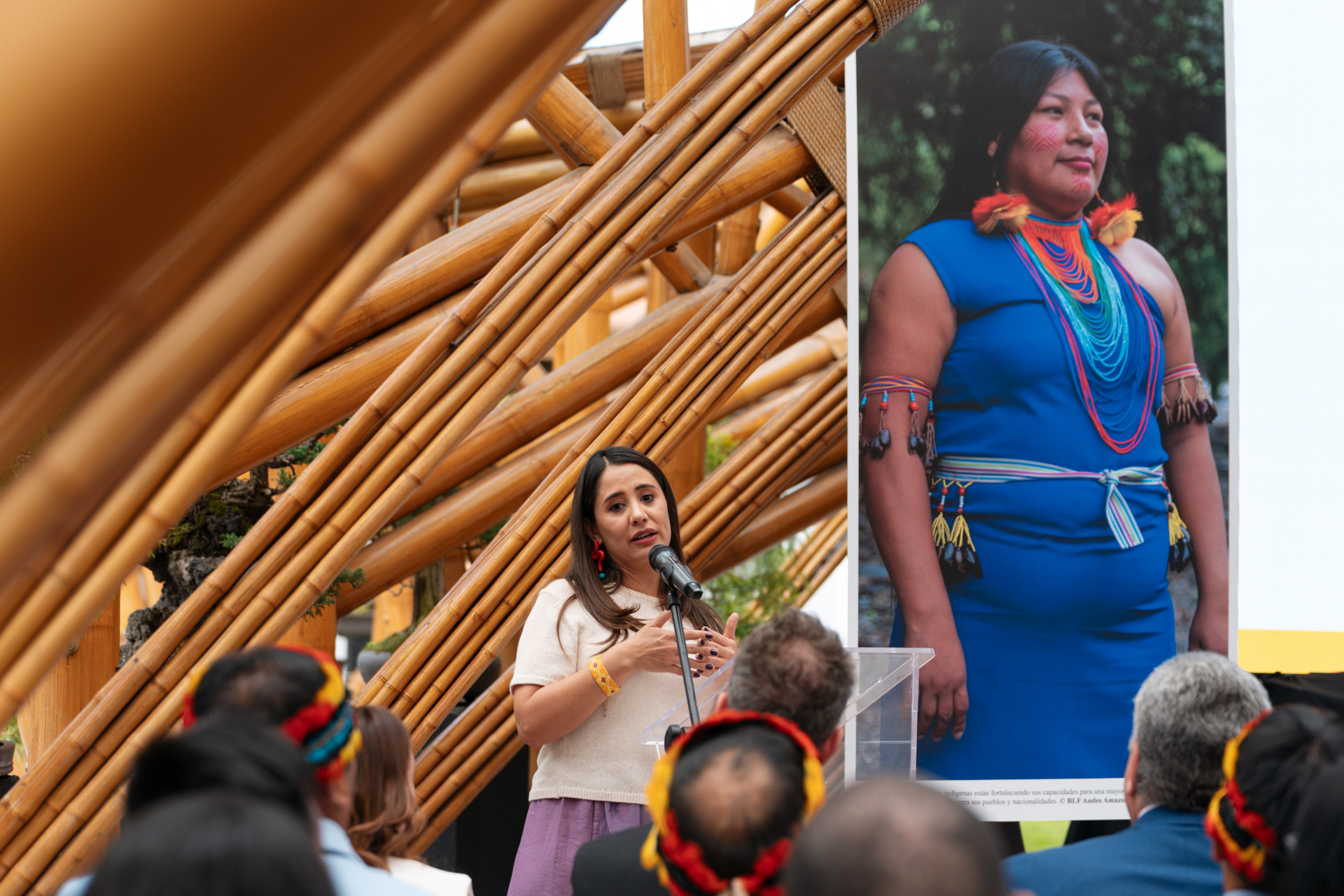A woman speaks at a podium in front of an audience, beside a large photo of an indigenous woman wearing traditional attire, in a bamboo structure setting.