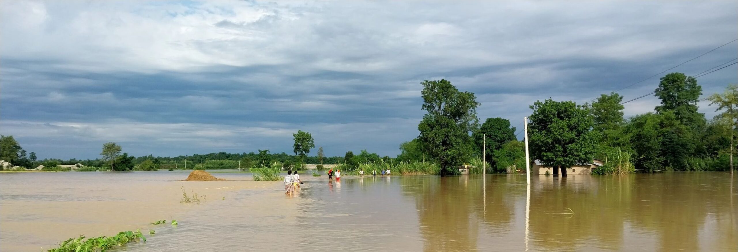 People walk through a flooded rural area with trees and submerged structures under a cloudy sky.