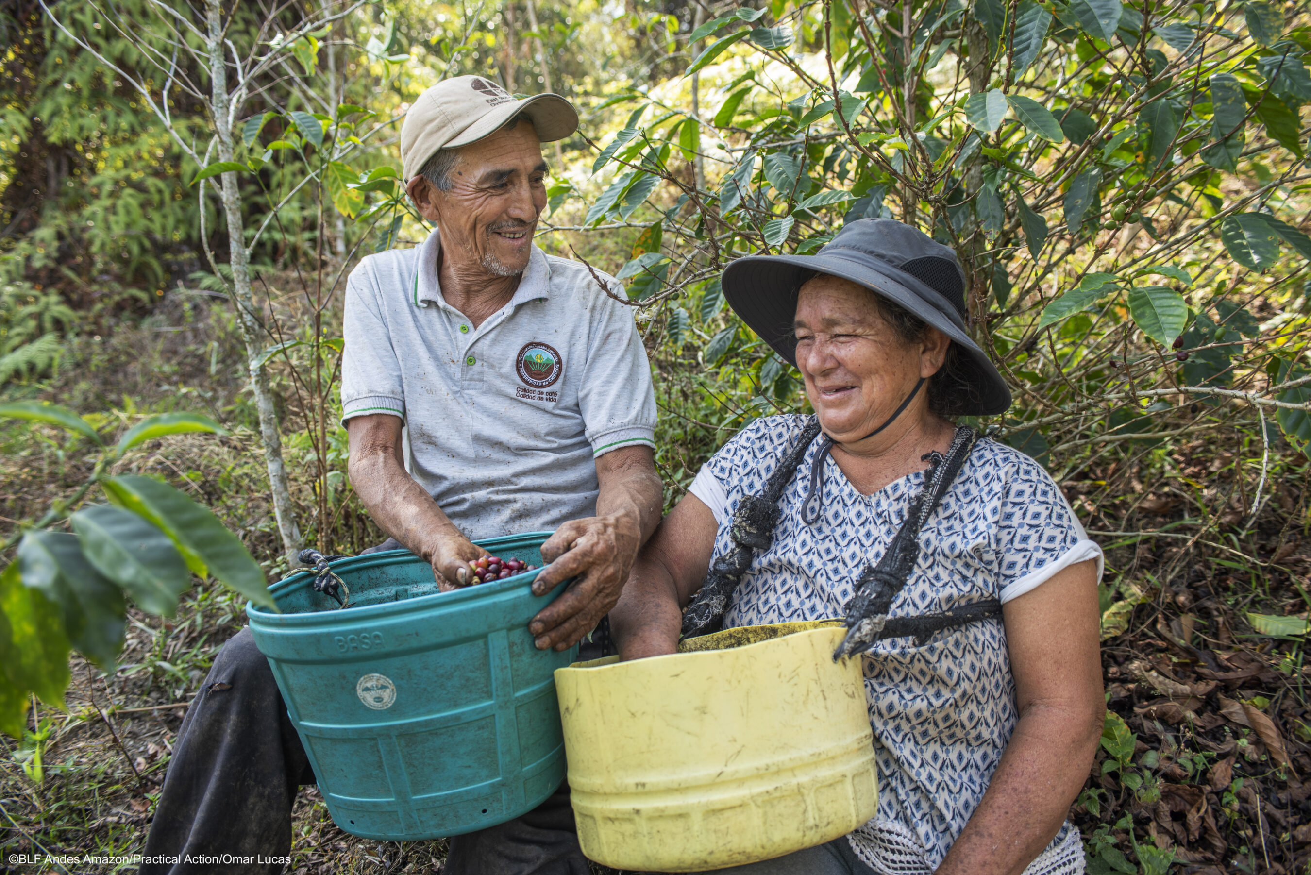 Two people sit outdoors among plants, smiling and holding buckets filled with harvested berries or fruit.