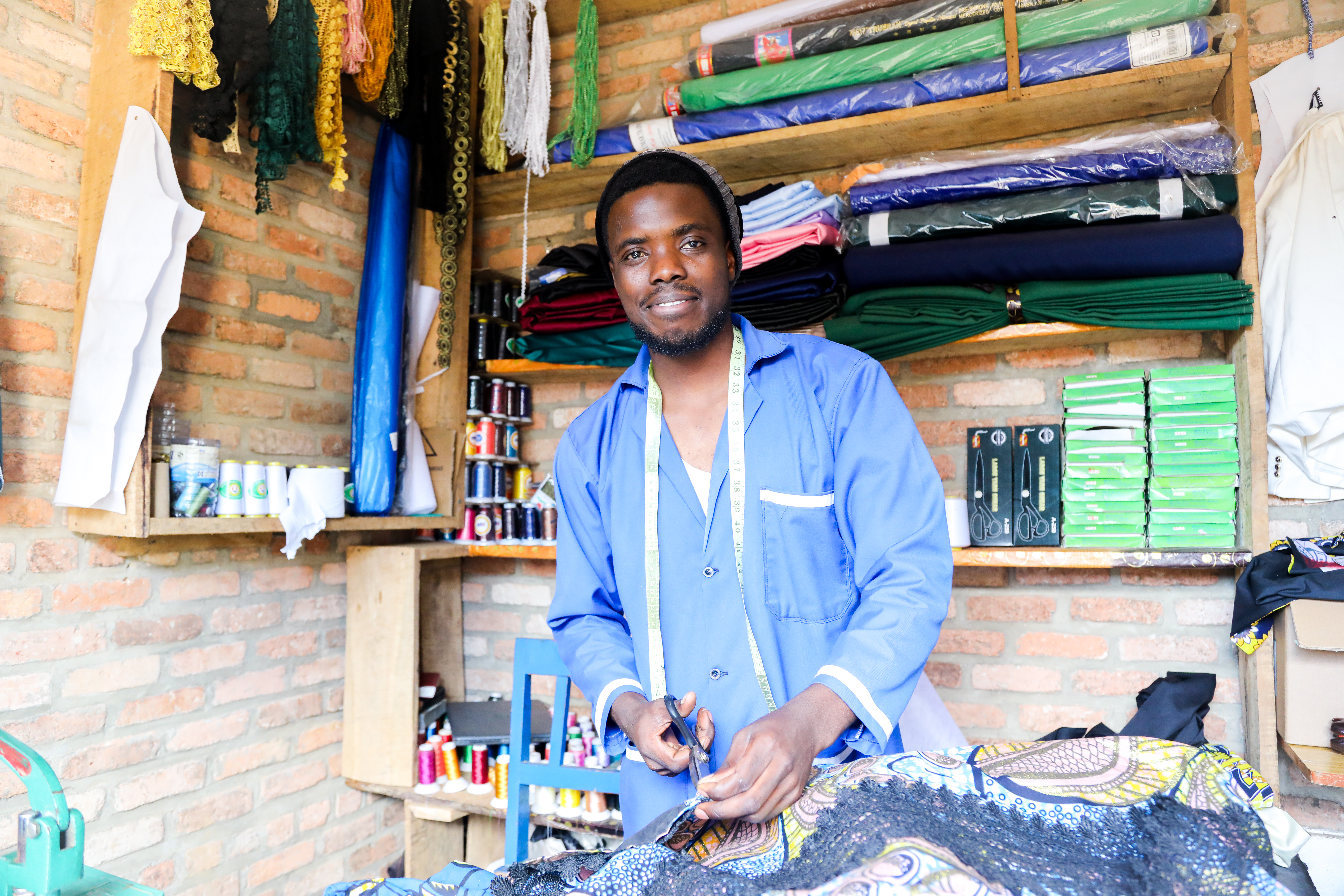 A tailor in a blue coat stands in a shop with fabric, thread, and shelves of supplies, holding scissors and working on patterned cloth.