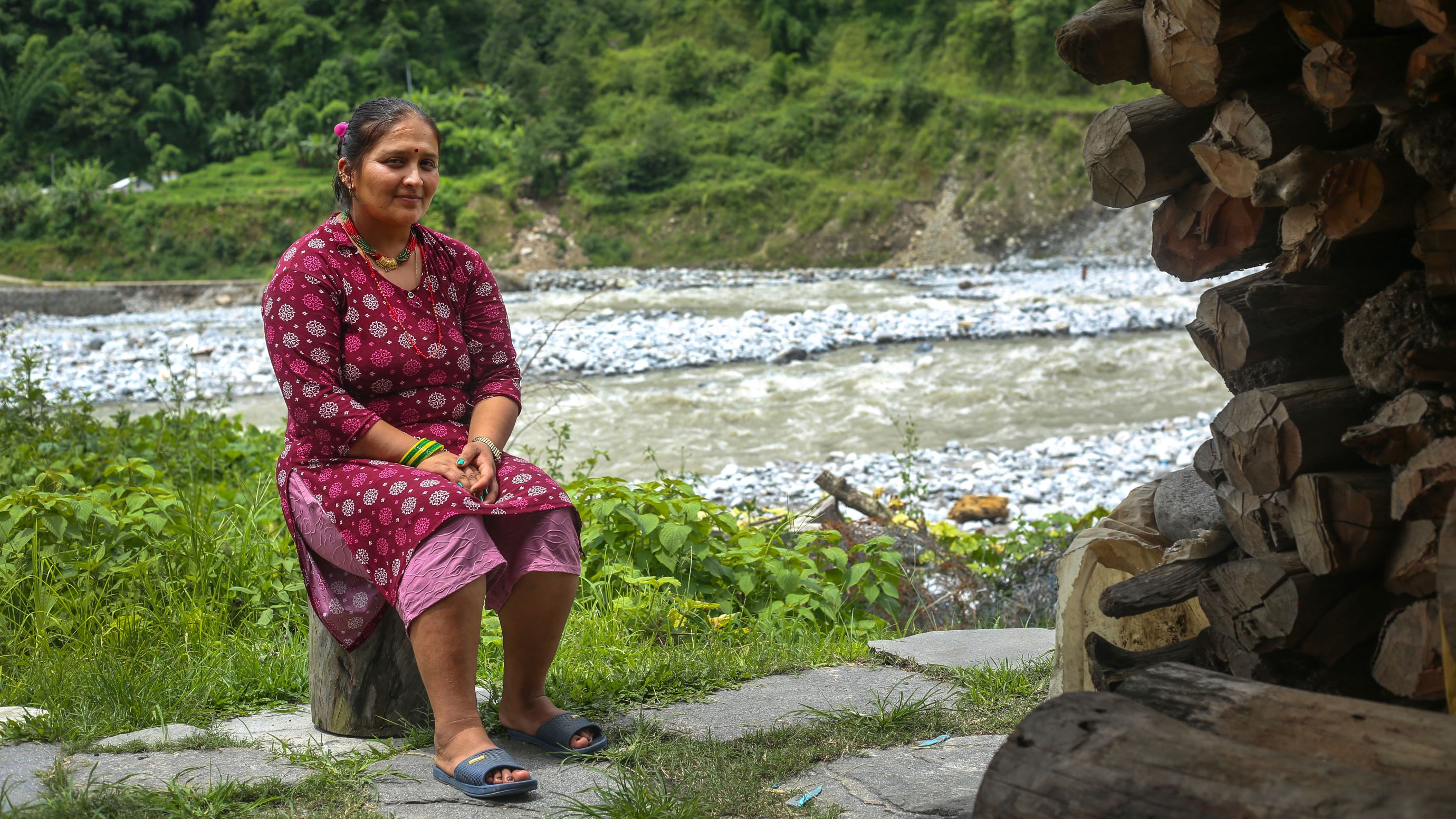 A woman in a red patterned dress sits on a log outdoors near a river, with stacked firewood and greenery in the background.