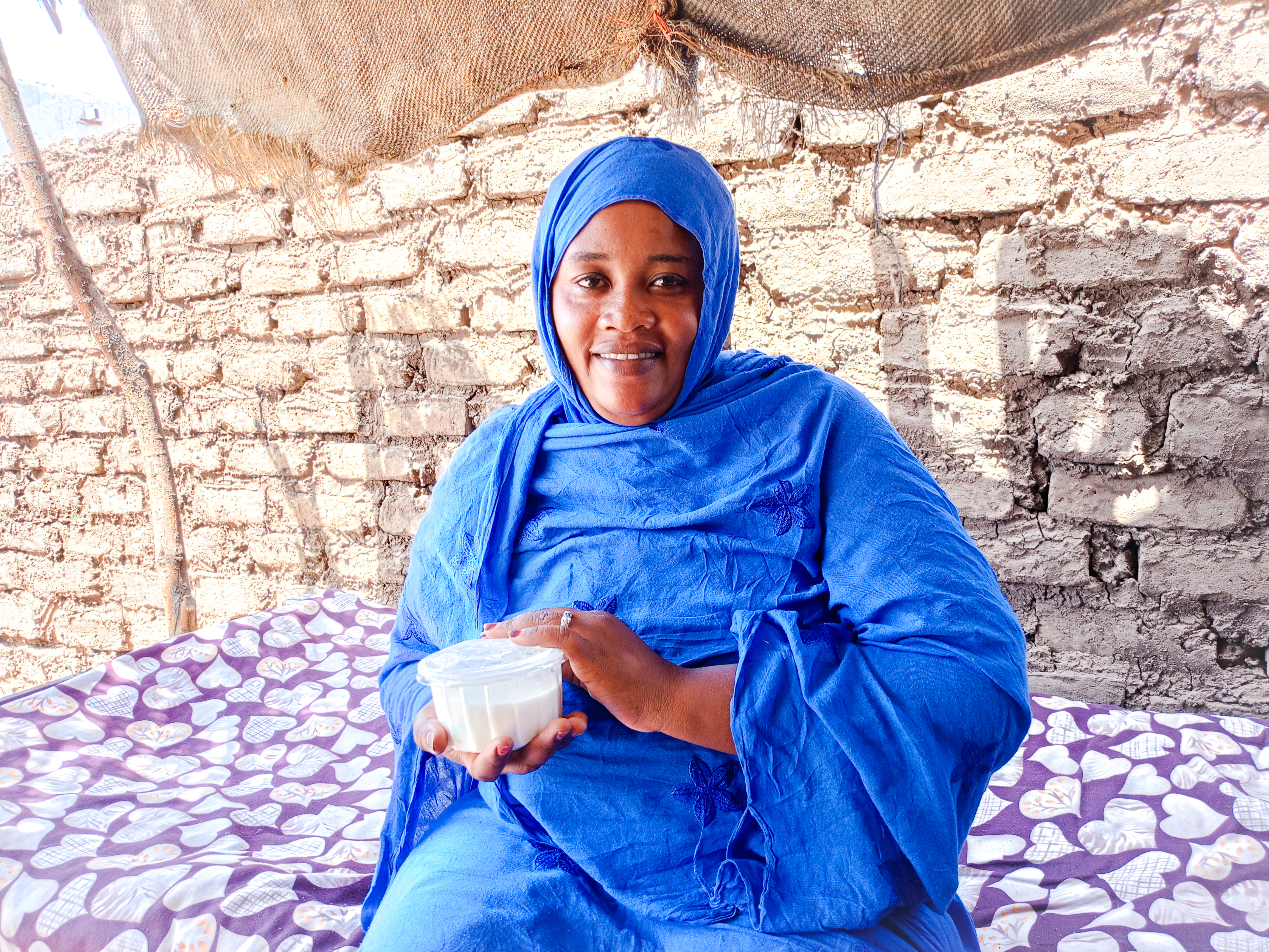 A woman wearing a bright blue dress and headscarf sits on a patterned bed, smiling and holding a plastic container of food against a background of stone walls.