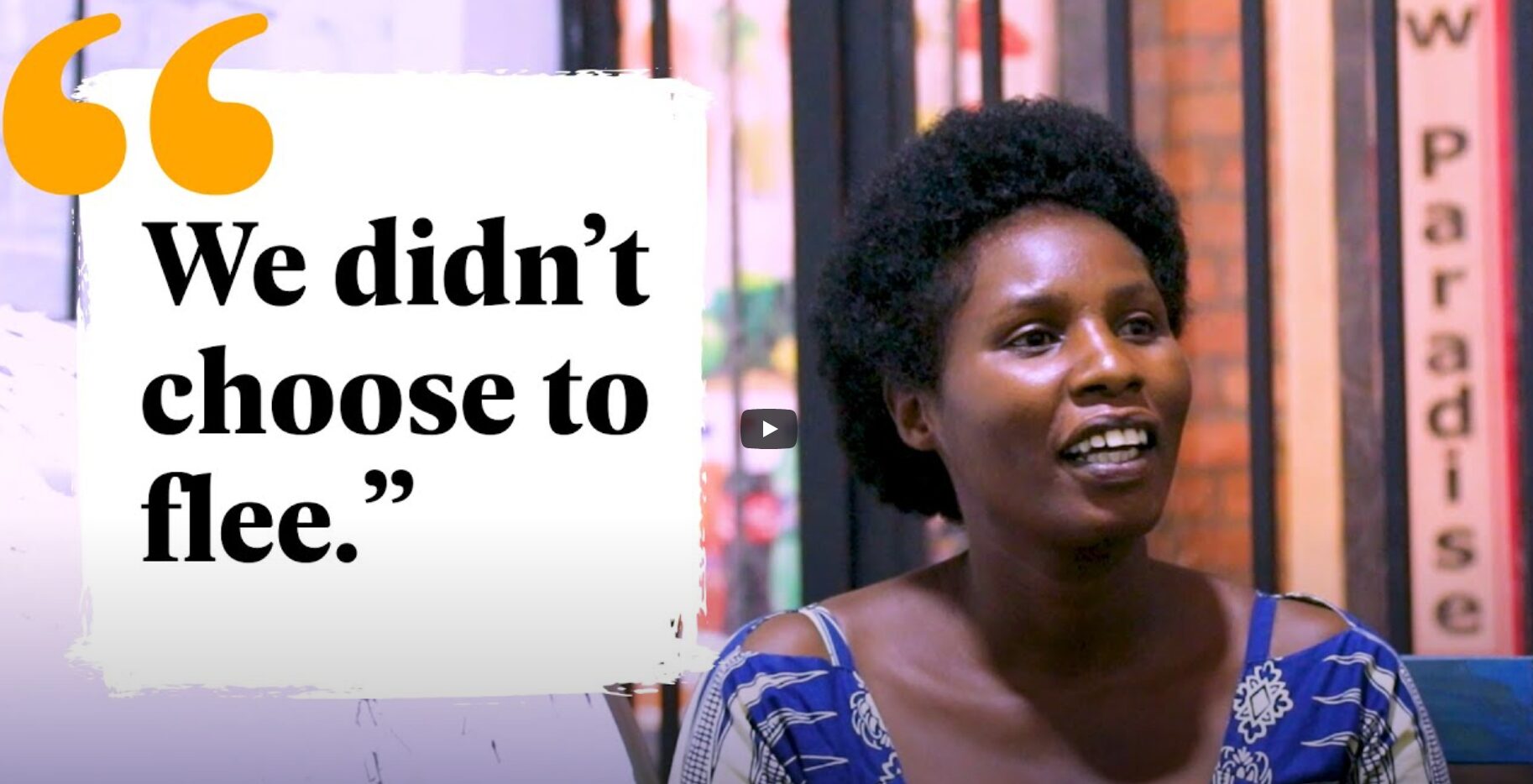 A woman with short curly hair sits and smiles next to a large quote reading, “We didn’t choose to flee.” Vertical signs with text are in the background.