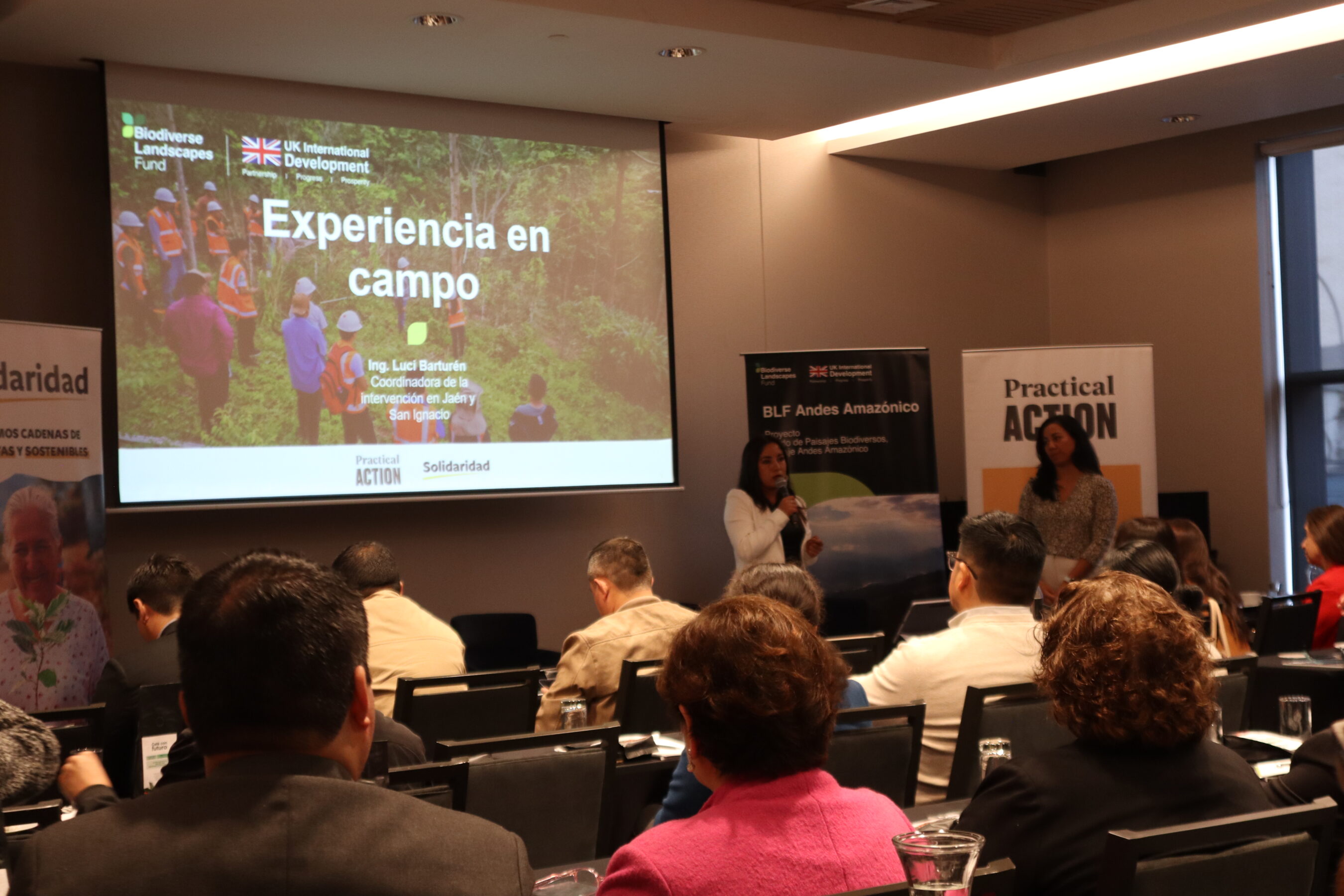 A woman speaks to an audience in a conference room with a presentation slide titled "Experiencia en campo" displayed on a screen behind her.