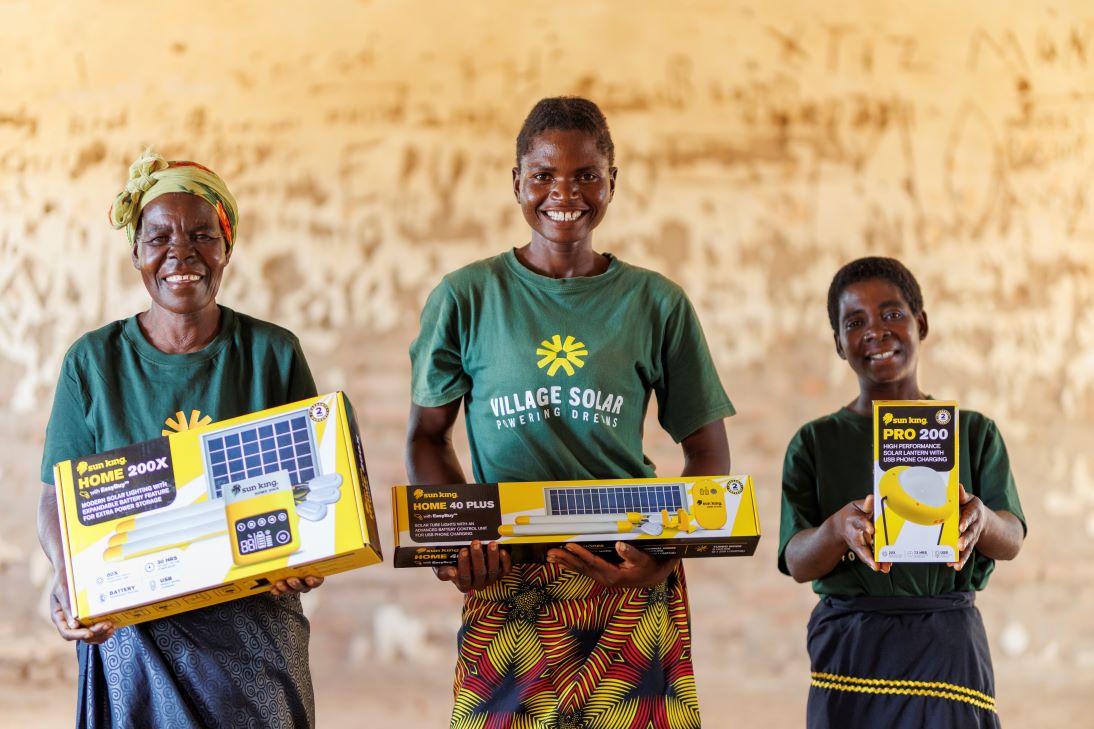 Three smiling women stand indoors holding solar power kit boxes, wearing green