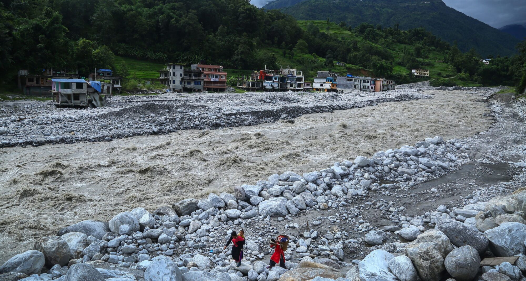 Two people walk across rocks near a swollen, fast-flowing river with houses and green hills in the background.