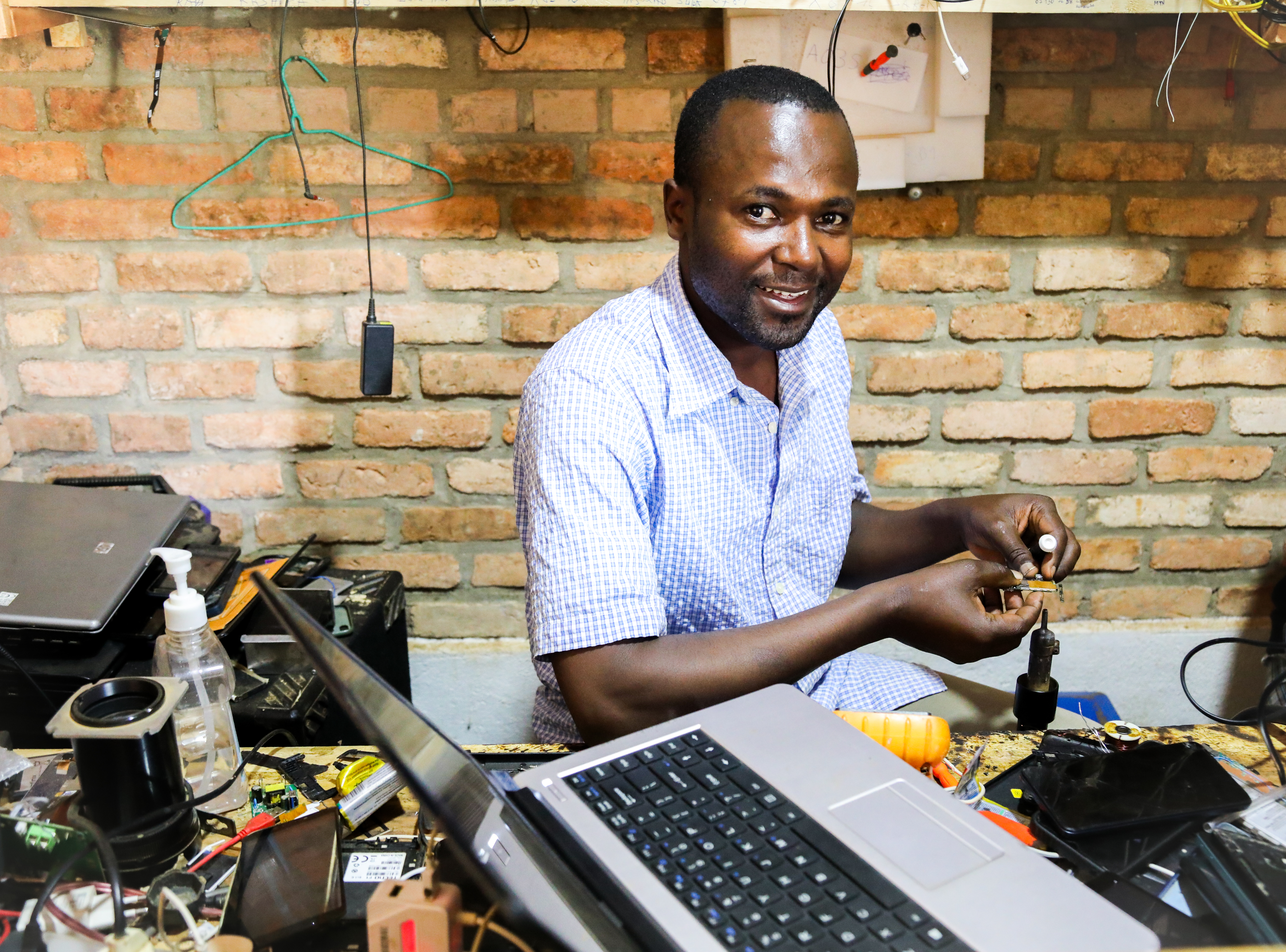 A man sits at a cluttered workstation with electronics, tools, and a laptop, smiling at the camera while working on a small device.