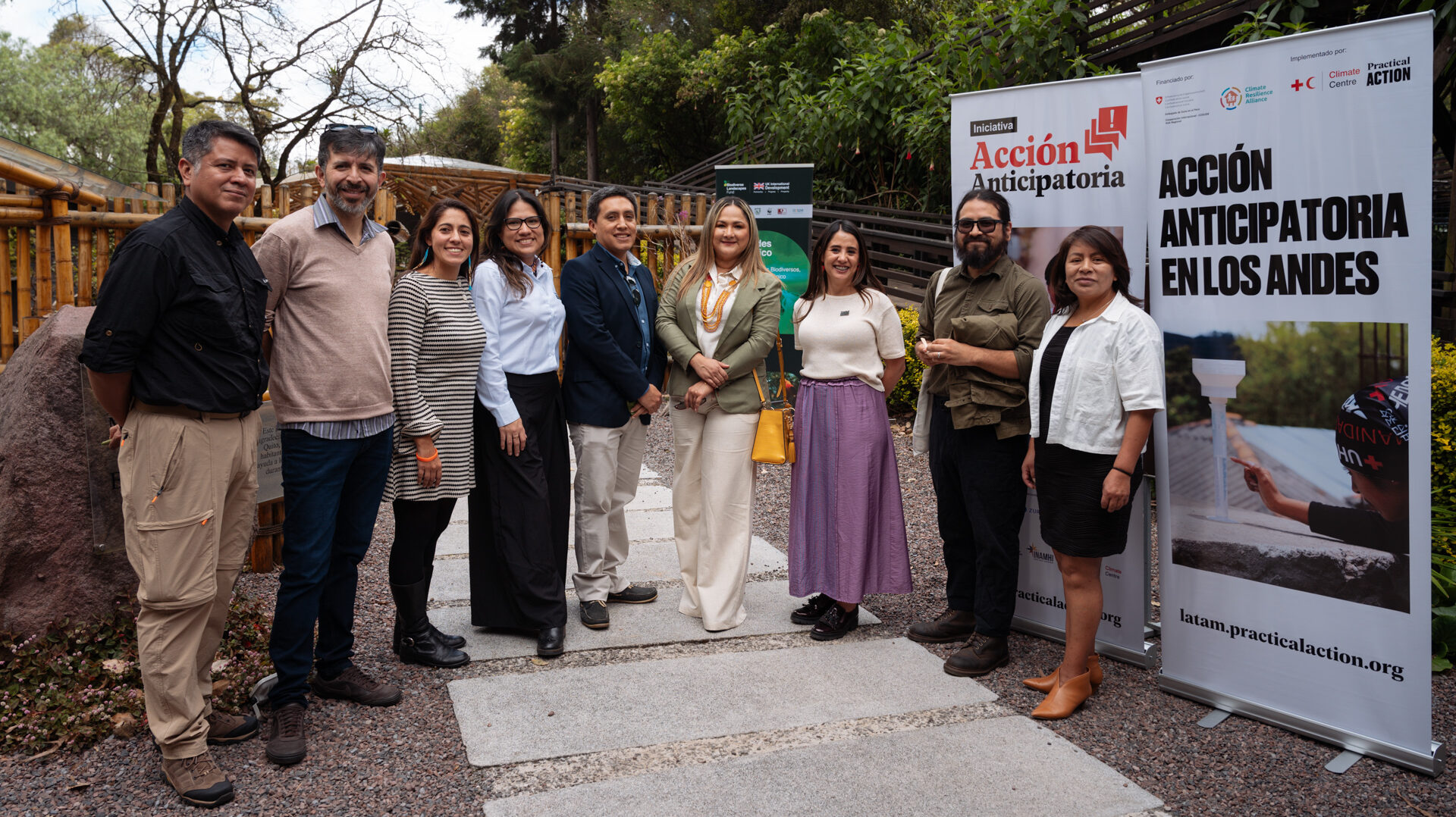 A group of ten people stands outdoors in front of banners for Acción Anticipatoria en los Andes, posing for a group photo on a stone path surrounded by greenery.