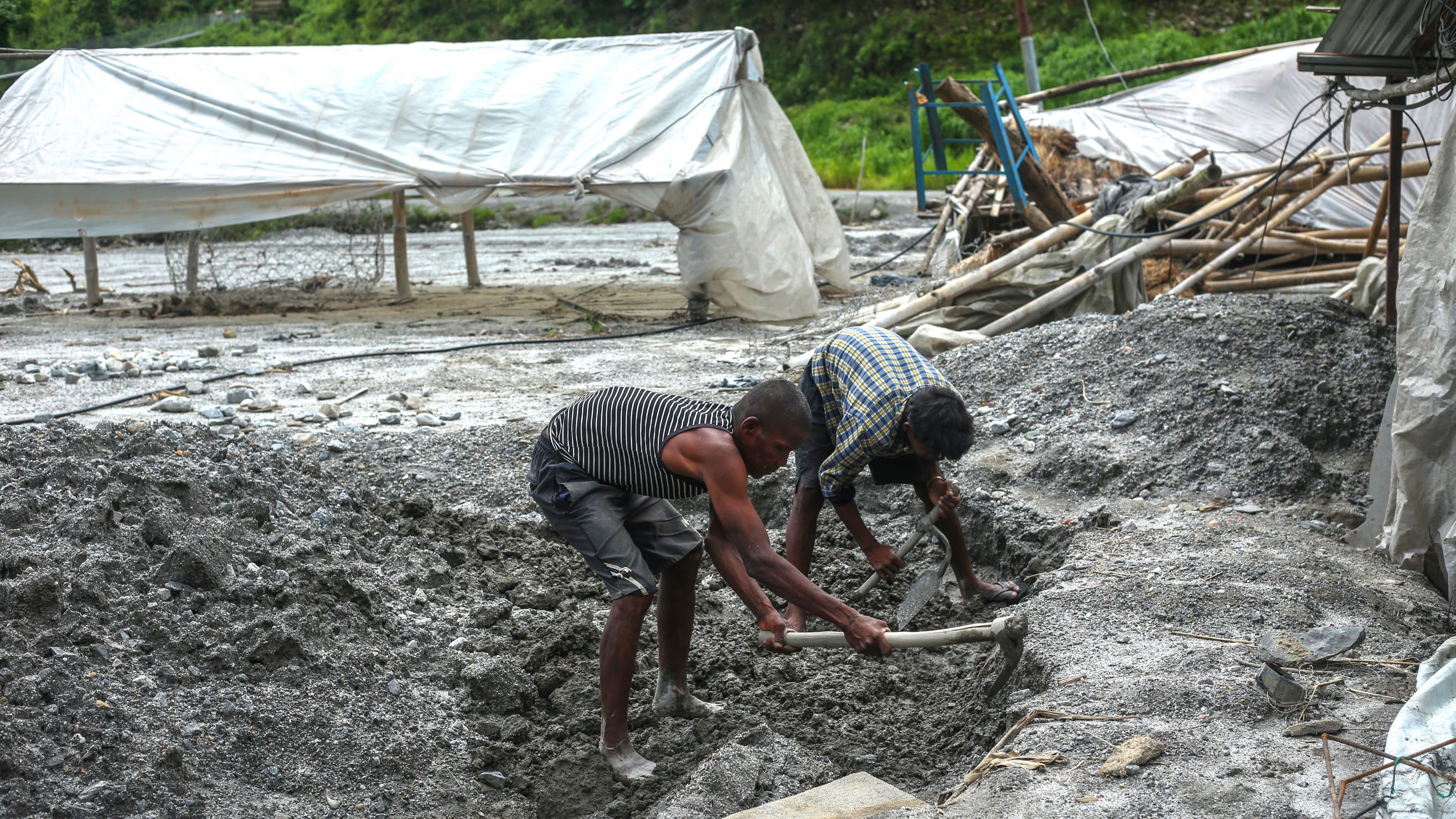 Two people work in a muddy outdoor area, using shovels to dig into the ground near a makeshift tent covered with white tarps.
