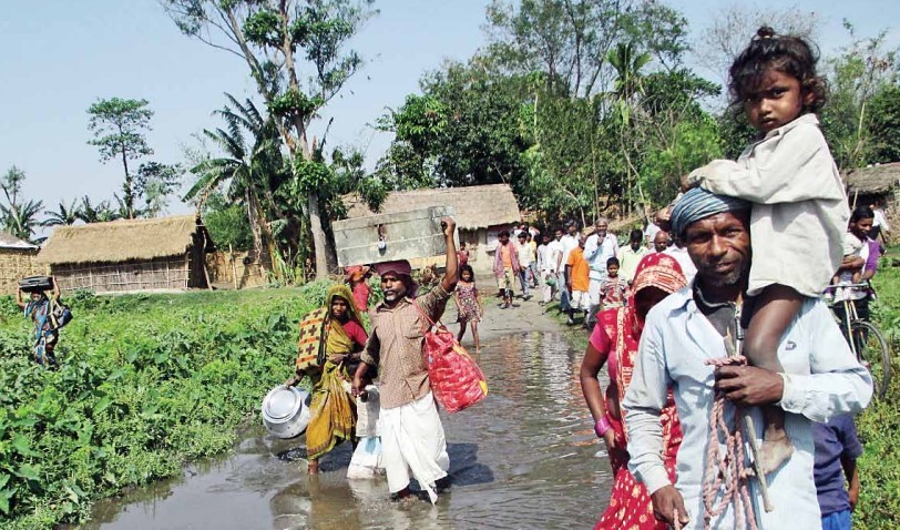 A group of villagers, some carrying belongings and children, walk through floodwater on a rural path with houses and greenery in the background.