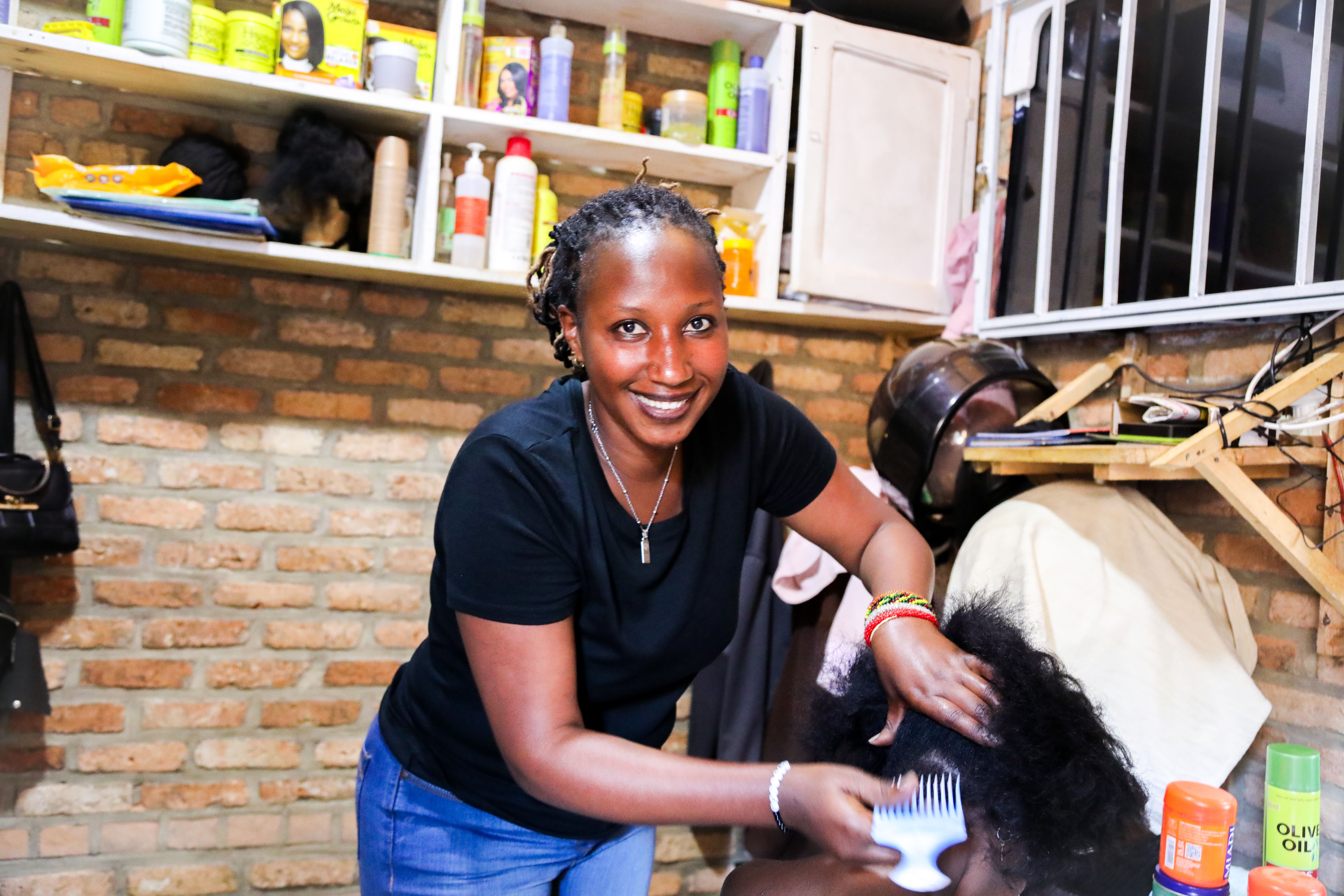 A woman smiling while styling another person’s hair in a salon with hair products on shelves and brick walls in the background.