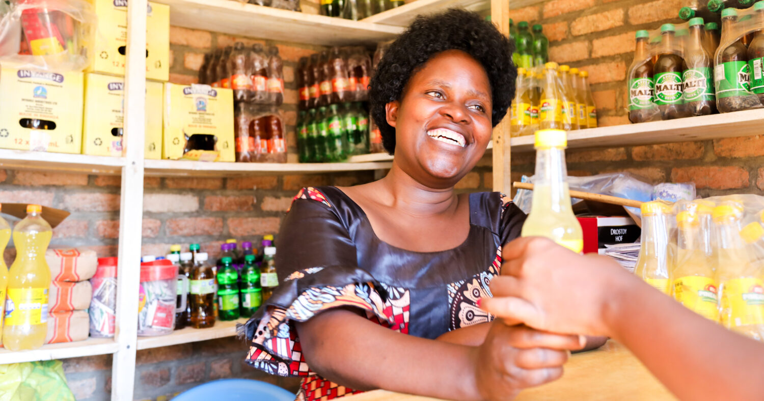 A woman smiles behind a counter in a small shop, handing a bottle of yellow drink to a customer. Shelves behind her are stocked with various drinks and packaged goods.