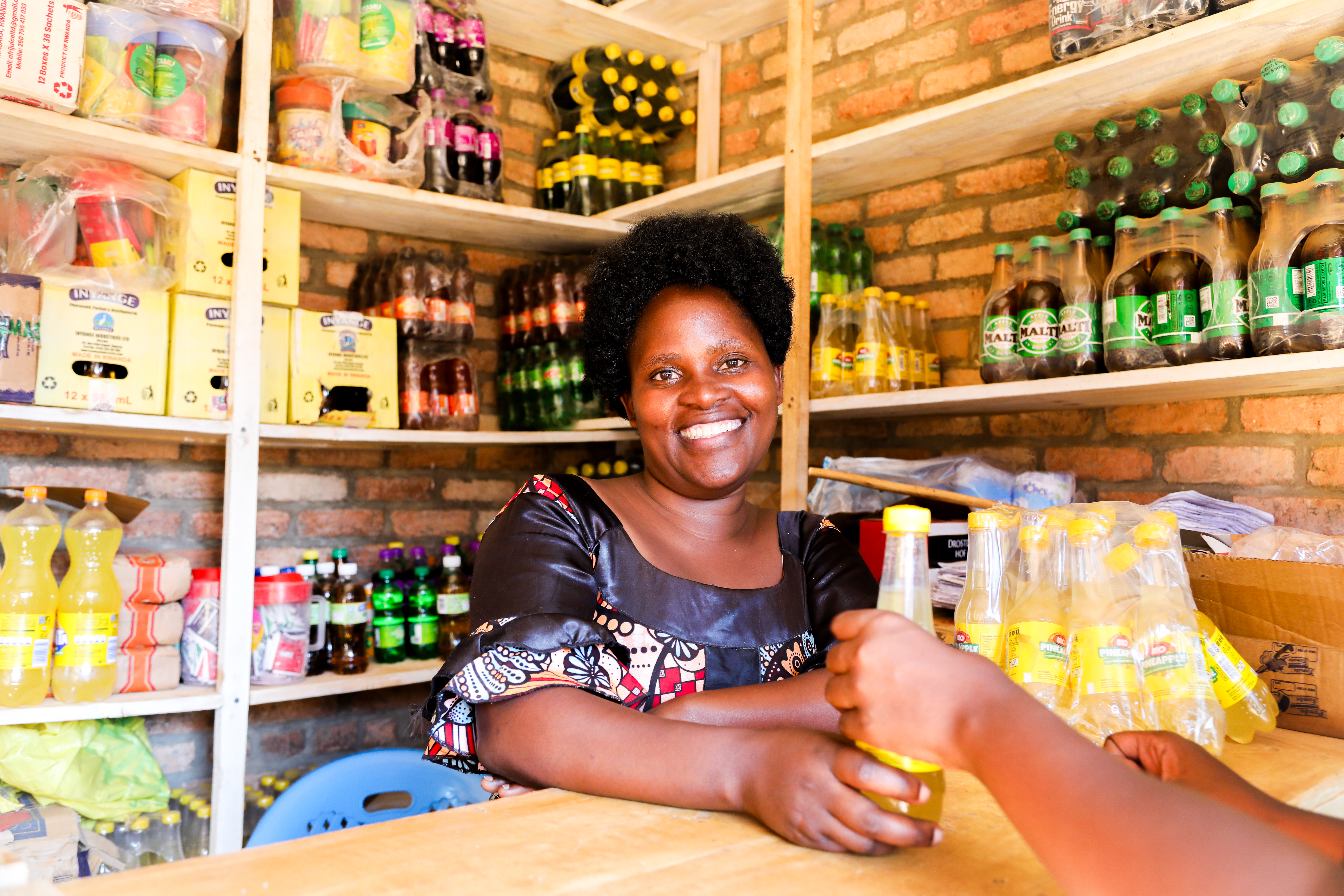 A woman stands behind a small shop counter, smiling, while handing a bottle to a customer. Shelves behind her display various bottles and packaged drinks.
