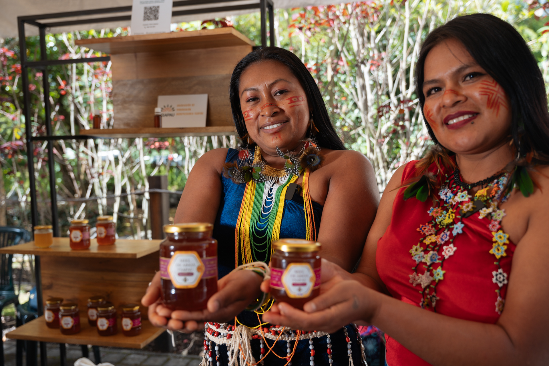 Two women in traditional attire with face paint hold jars of honey, standing in front of a display shelf with more jars and a QR code in an outdoor setting.