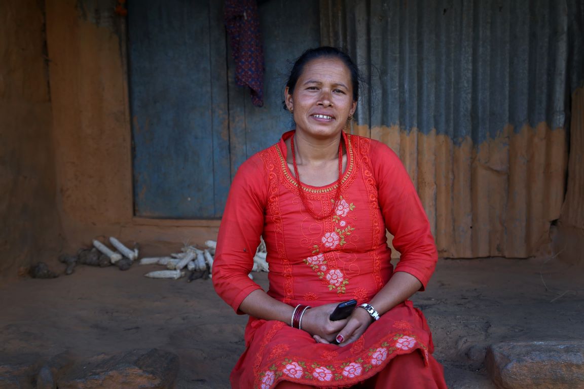 A woman in a red dress sits outdoors on a dirt surface, holding a phone, with a corrugated metal and wooden wall in the background.
