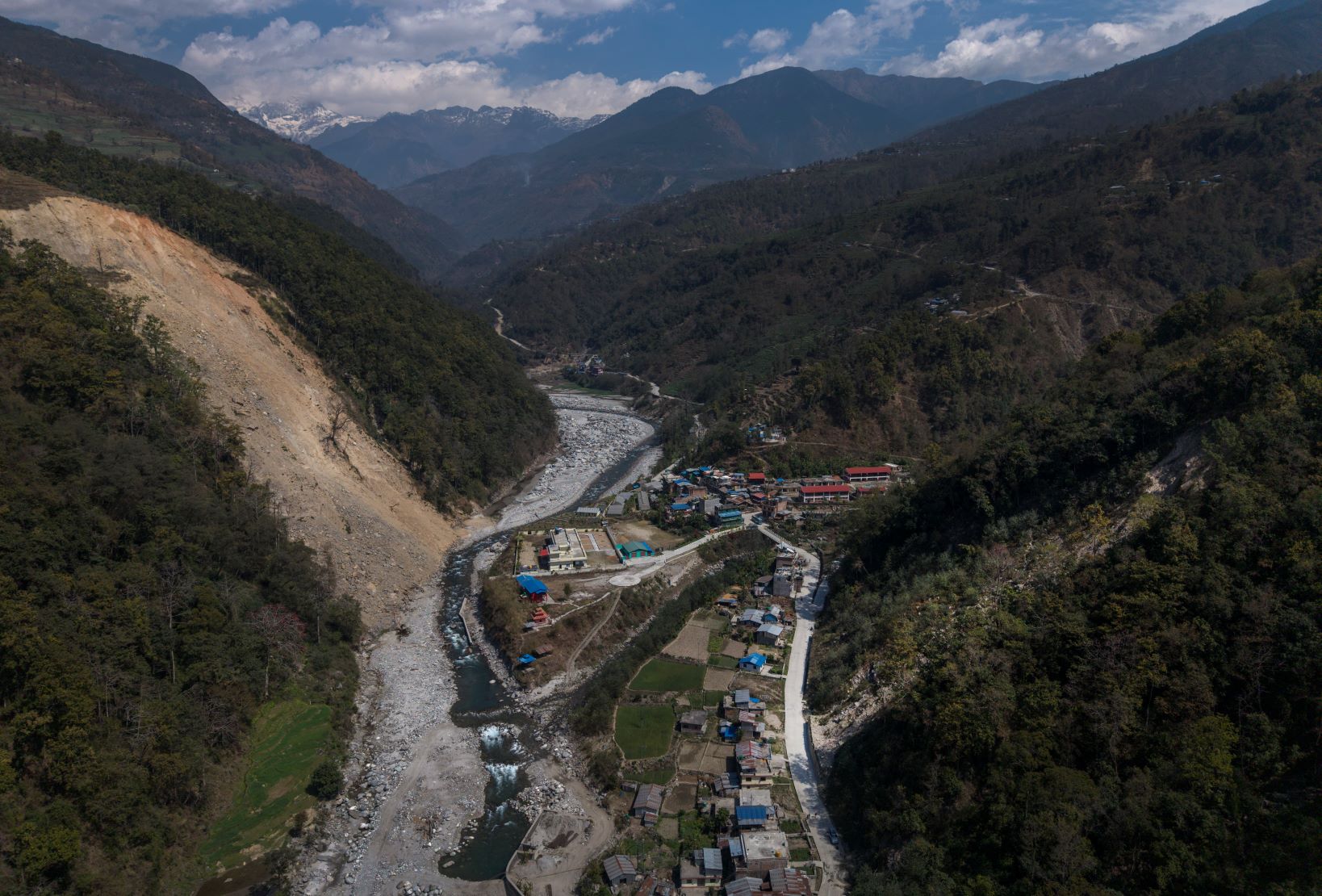 A river runs through a mountain valley with scattered buildings on both banks, surrounded by forested hills and distant peaks under a partly cloudy sky.