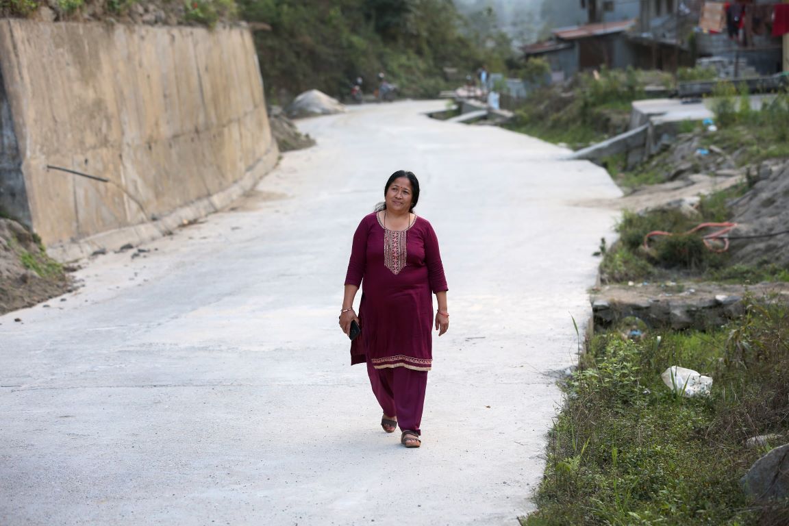 A woman in a maroon outfit walks alone on a concrete road bordered by greenery and a steep hillside.