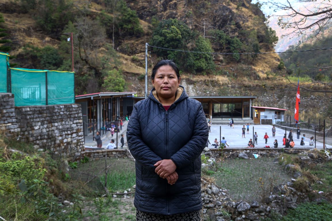 A woman in a dark quilted jacket stands outdoors in front of a stone wall and a group of people gathered near a building in a mountainous area.